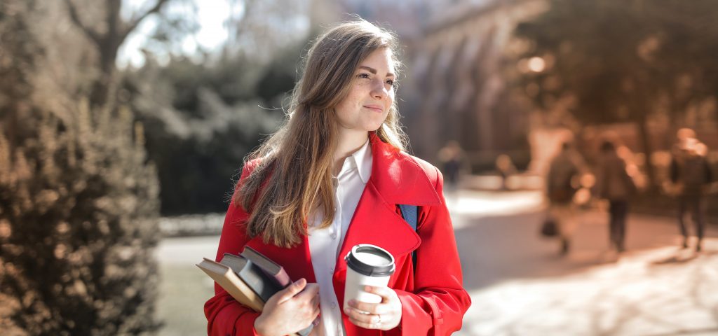 Woman wearing red coat, holding book and coffee as she walsk through university grounds