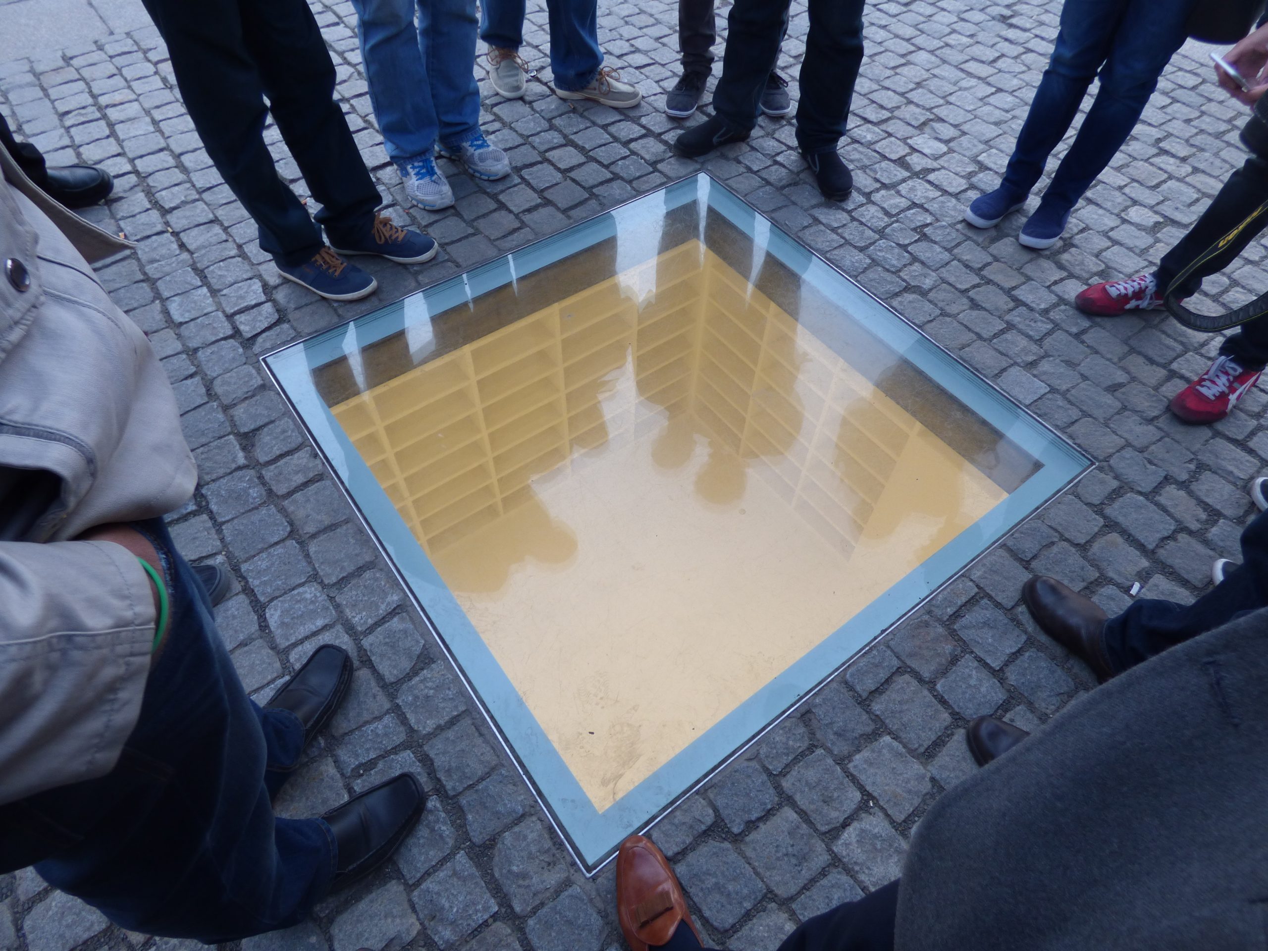 Photo of people feet in the street looking at a monument in a the ground that is a selection of empty bookcases,