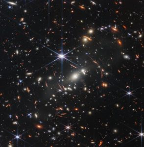 Thousands of small galaxies appear across this view. Their colours vary. Some are shades of orange, while others are white. Most appear as fuzzy ovals, but a few have distinct spiral arms. In front of the galaxies are several foreground stars. Most appear blue, and the bright stars have diffraction spikes, forming an eight-pointed star shape. There are also many thin, long, orange arcs that curve around the centre of the image.