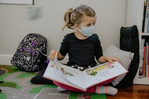 Child sitting cross-legged reading a picture book. She is wearing a mask.