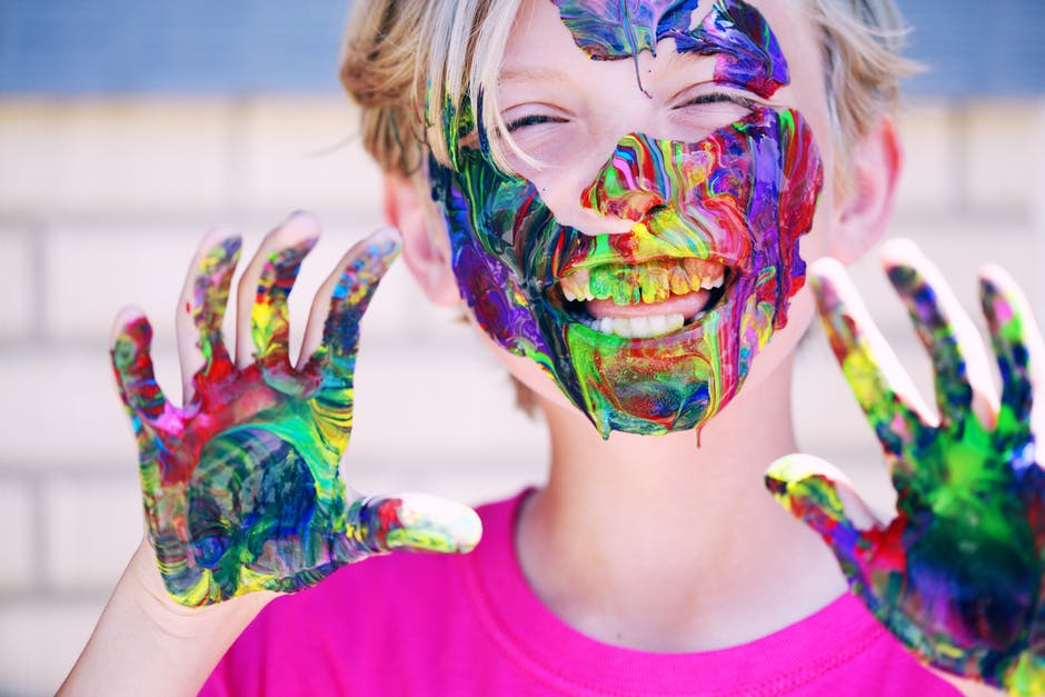 Child who's face and hands are covered in colourful paint. They are smiling with joy.