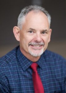 White middle aged man with greying hair and beard. Wearing blue checked shirt and red tie