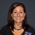 Middle-aged woman with tanned skin and shoulder length dark brown hair. She is smiling