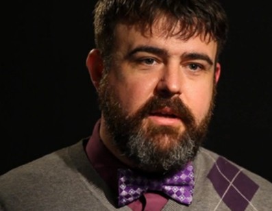 Mr Erik Gordon, a white male with a dark brown beards and hair, wearing a collared shirt, bow tie, and jumper.