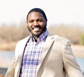 Mr James Moffatt, a black man with black hair and beard, dressed in a checked collared shirt and white suit jacket.